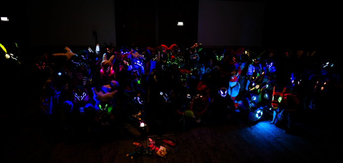 A group of protogen and synth fursuiters sitting in the dark, illuminating only from their LEDs