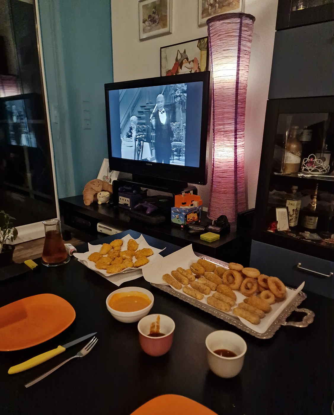 Photo of a living room with Dinner for One running on an old flatscreen TV, while an opulent banquet is set on the coffee table. Wide ranging treats from faraway countries, such as mozzarella sticks, chicken nuggets, onion rings, and cream cheese jalapeños, along with a choice of dips.