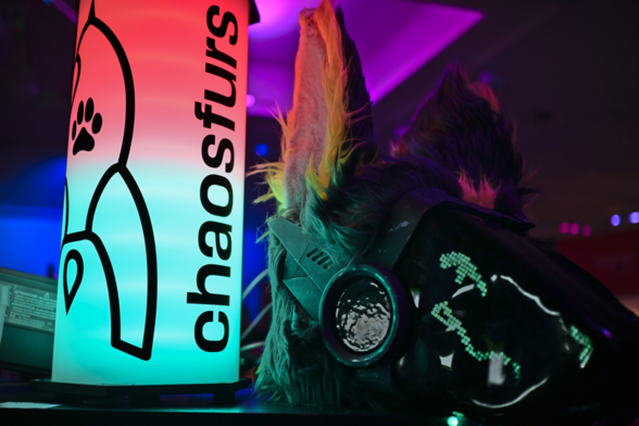 A protogen fursuit head with LED eyes and mouth next to a colorful lamp with "chaosfurs" and a fairydust rocket on it