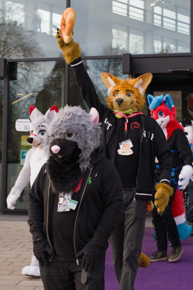 Fursuiters walking out of a building.

A grey rat in front, behind it an orange cat holding a croissant. Some more fursuits in the background