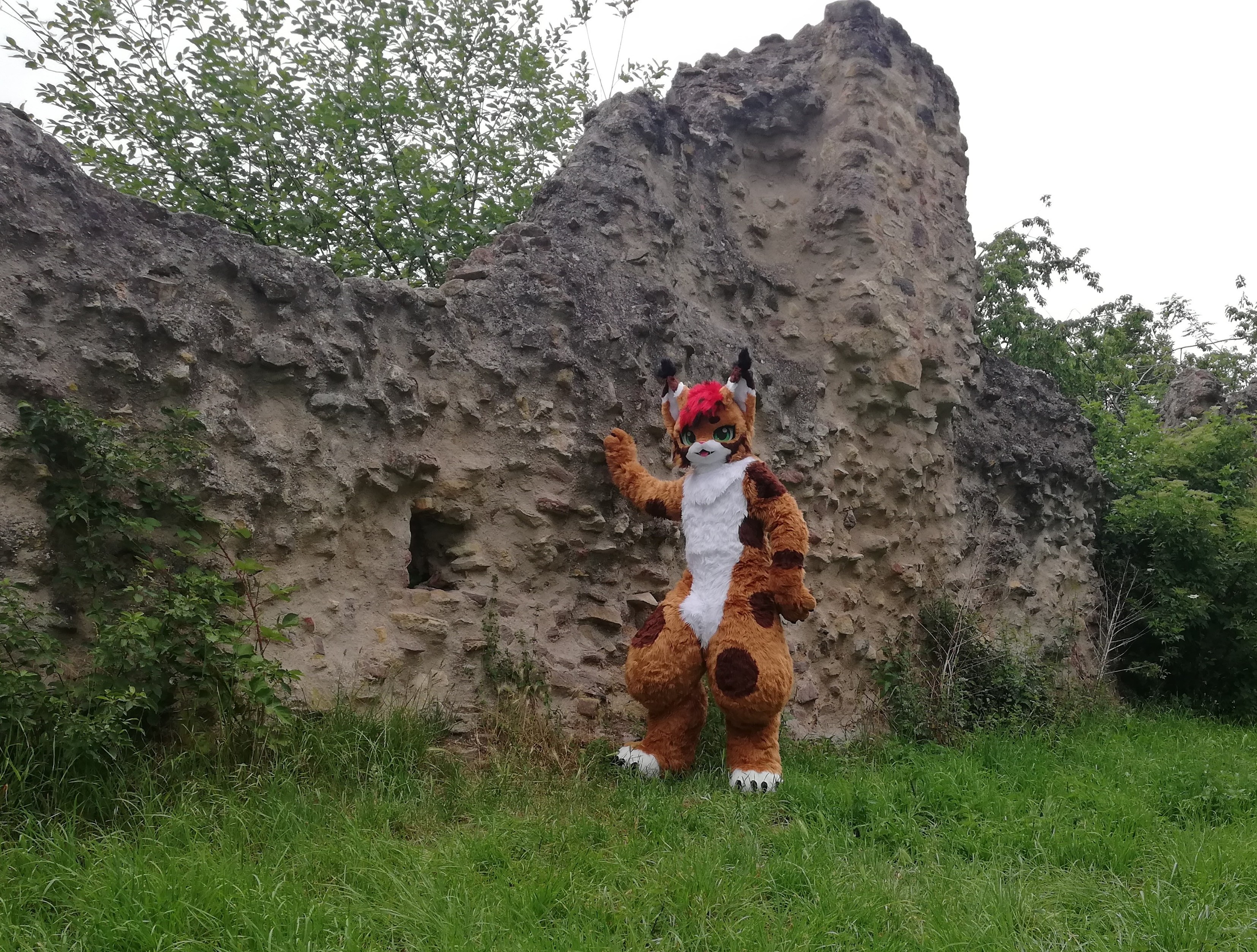 A brown and white lynx fursuiter with red hair and green eyes leaning against an ancient, broken brick wall
