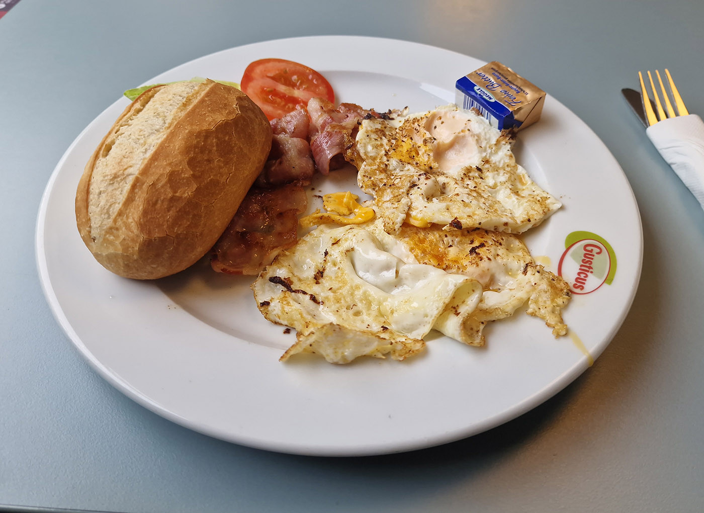 Photo of eggs and bacon with a bread roll and butter. The eggs were supposed to be sunny side up, but I guess they had some gruesome accident.