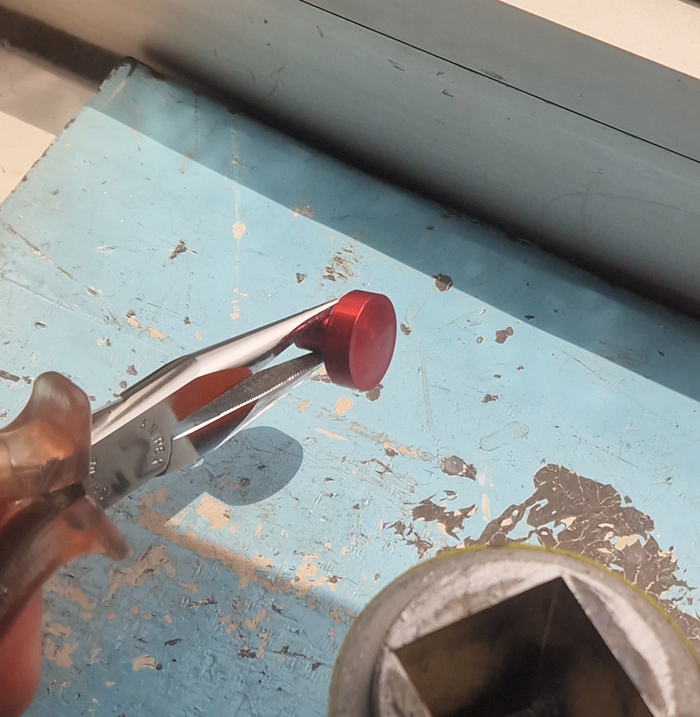 A photo of the fruits of all this labor: A small, red, cylindrical, pedestal-shaped object being held with tweezers and photographed from behind a lead shield through a lead-glass window.