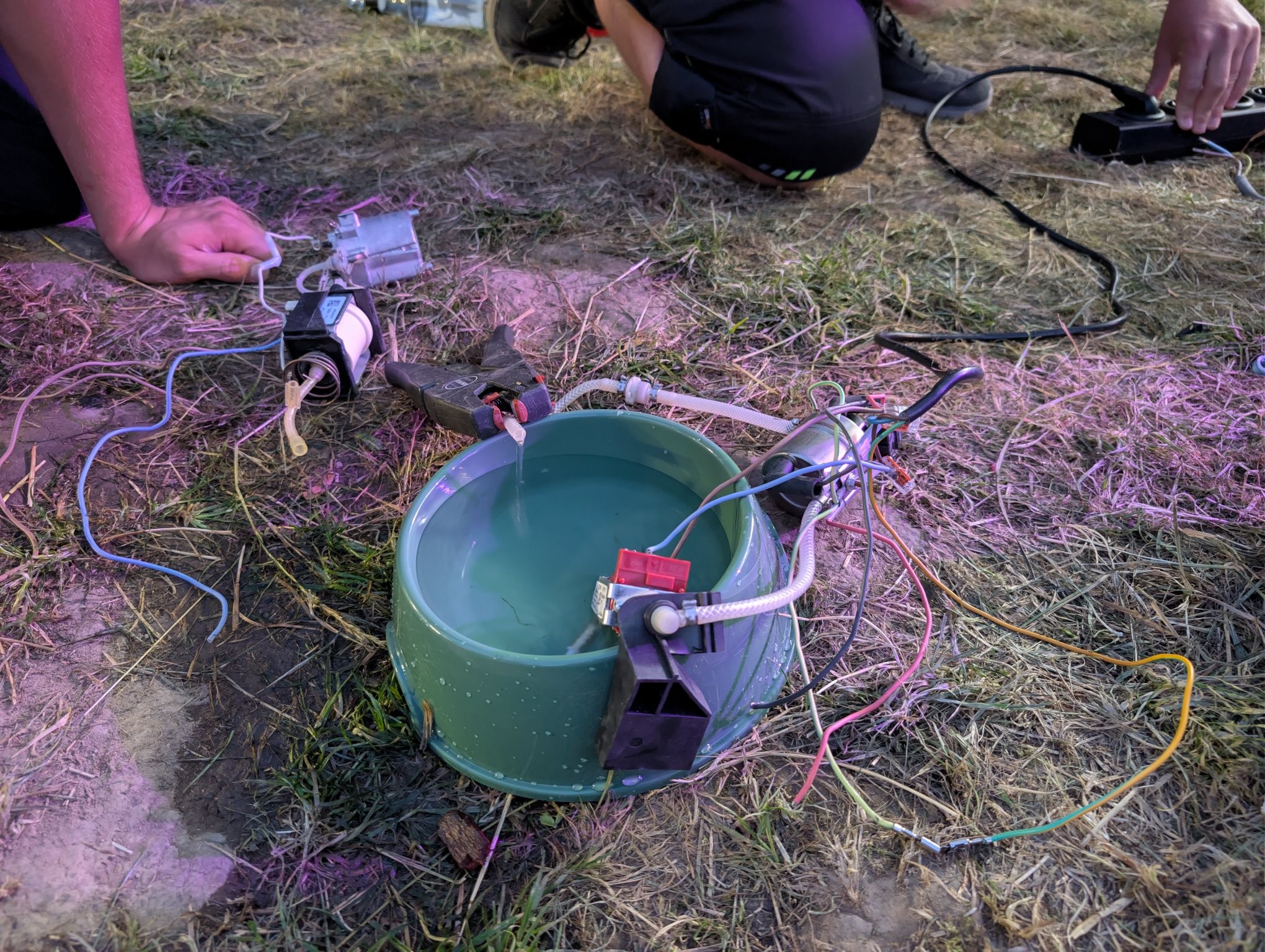 Ein grüner Hundenapf, in dem bzw. daneben eine Pumpe aus einer Kaffeemaschine und die dazugehörige Heizung liegen. Der Schlauch mit dem heißen Wasser geht zurück in den Napf. Alles ist lose und offen an 230V angeschlossen.