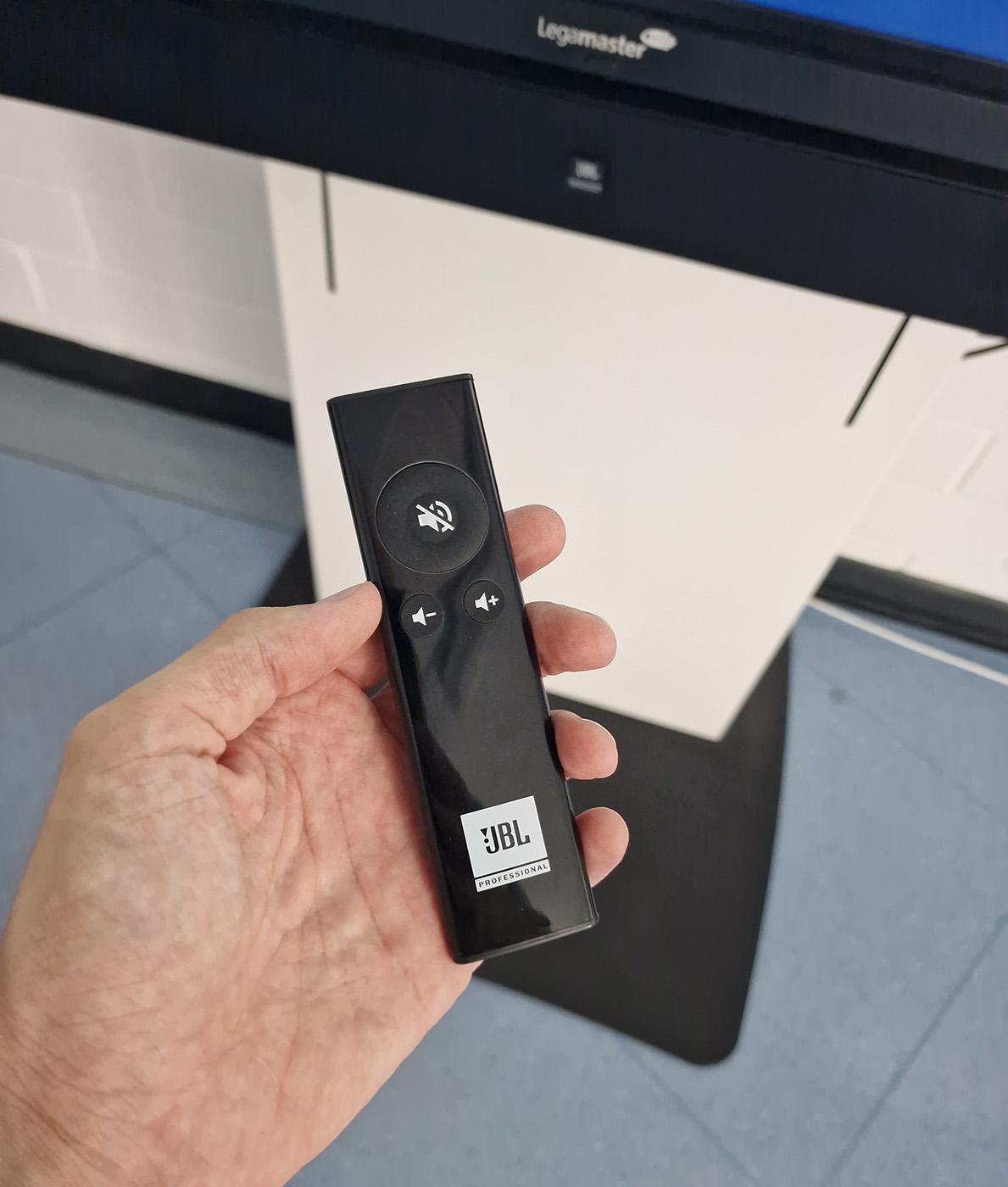 Photo of a hand holding a JBL remote for a sound bar of a large screen. It has been patiently sitting in its wrapper, in a box of miscellaneous surplus parts of the video system that had been set aside when it was all set up. The sound bars also had some controls at their back, allowing you to switch between a fixed and variable input source volume, and four levels of output. What confused me the most was that no matter how I set up the sound bar, it doesn't care at all about whether I have volume set to 0 or 100 under Windows or Zoom, which is a behavior my brain doesn't really comprehend that well. But now we have the remote for direct control, so that's okay.