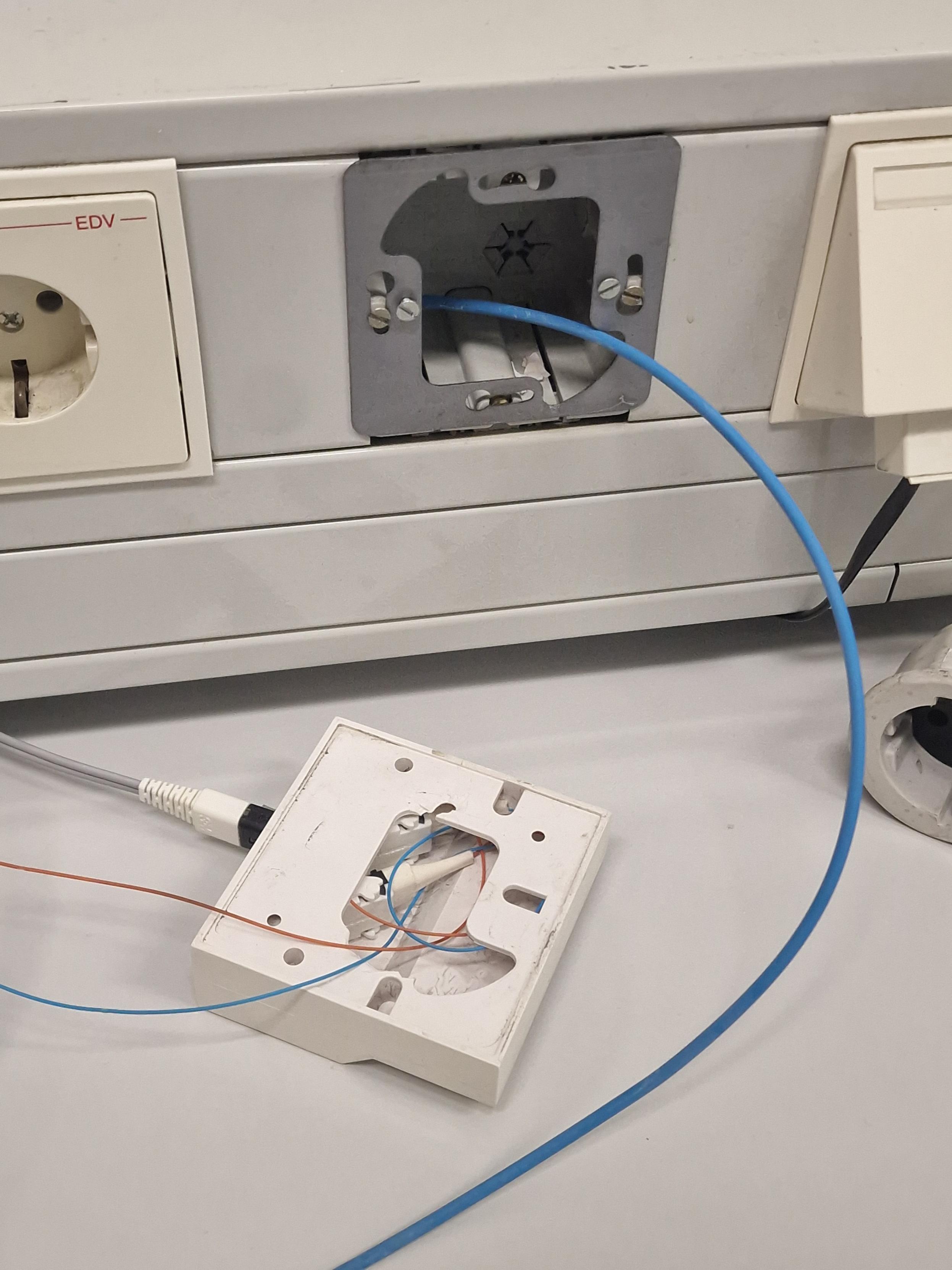 Photo of a cable duct, with an optical fiber coming out of a hole that is obviously intended for the junction box currently chilling on a table, due to the fact that the backplates where the screws would attach it to the wall have fractured.