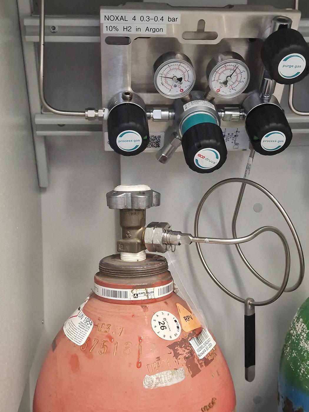 Photo of the top of a gas cylinder, connected to its pressure regulator inside a safety cabinet. Some droplets of water are visible from the condensation that had formed on the valve.